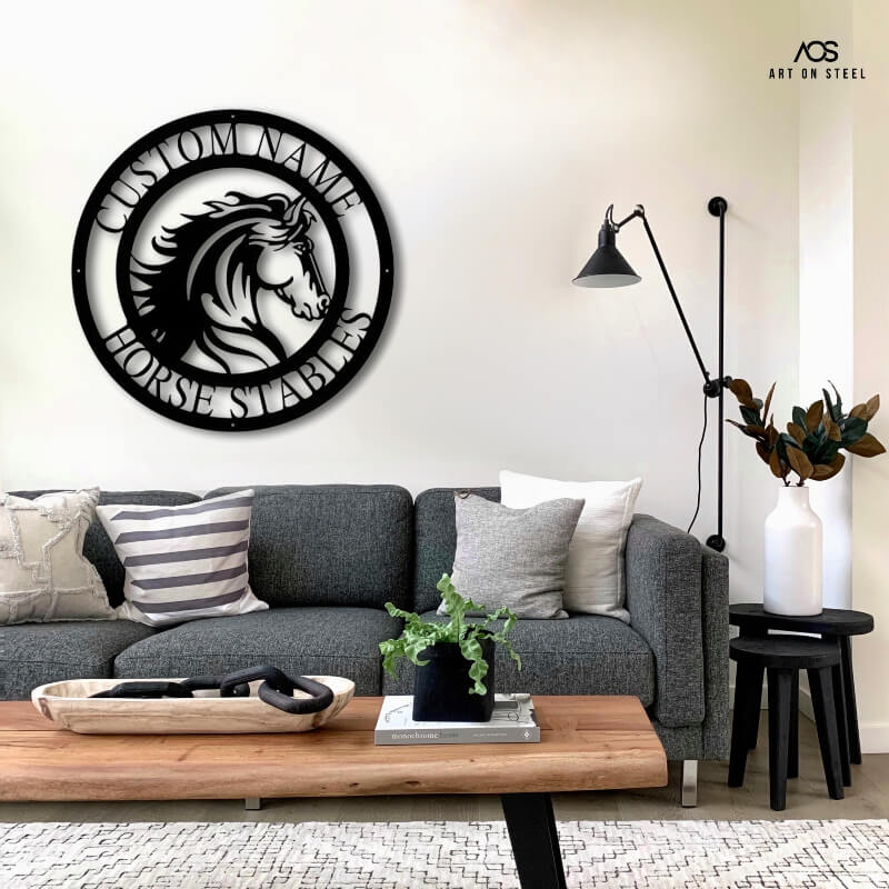 A circular black metal sign featuring a stylized horse head with a flowing mane in the center. The text "CUSTOM NAME" and "HORSE STABLES" is written around the border. The sign is mounted on a light-colored wall above a gray couch in a modern living room, with a live-edge coffee table, a black lamp, and a white vase. The "Art on Steel" logo is visible in the top right corner.