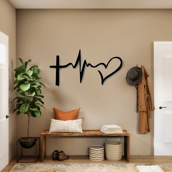 A close-up of a modern, black steel wall art piece featuring a cross, a heartbeat line, and a heart, symbolizing faith, hope, and love. The art hangs above a rustic wooden bench in a home entryway, which is decorated with a potted plant, a hat, a brown jacket, and woven baskets. Art on Steel