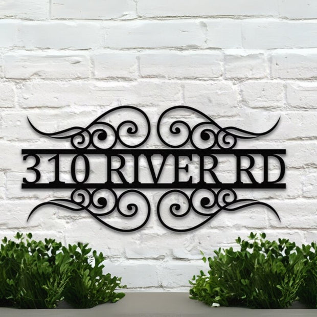 A custom, black metal house number sign featuring the number 310 and the street name RIVER RD framed by elegant wrought-iron scrollwork. The sign is mounted on a white painted brick wall, with green hedges visible at the bottom of the image. Art on Steel