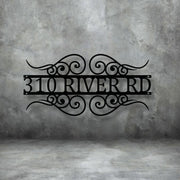 A close-up of a custom, house number sign, with the numbers 310 and the street name RIVER RD framed by decorative scrollwork. The black sign is mounted on a gray, distressed concrete wall. Art on Steel