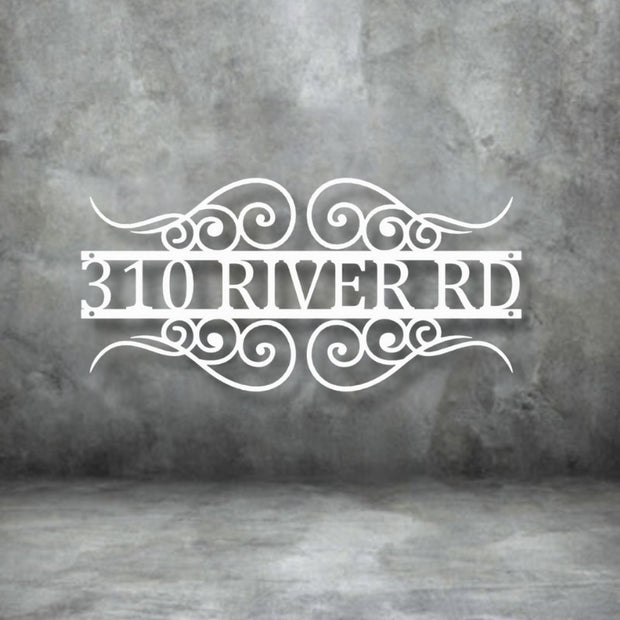 A custom, black metal house number sign featuring the number 310 and the street name RIVER RD framed by elegant wrought-iron scrollwork. The sign is mounted on a white painted brick wall, with green hedges visible at the bottom of the image. Art on Steel