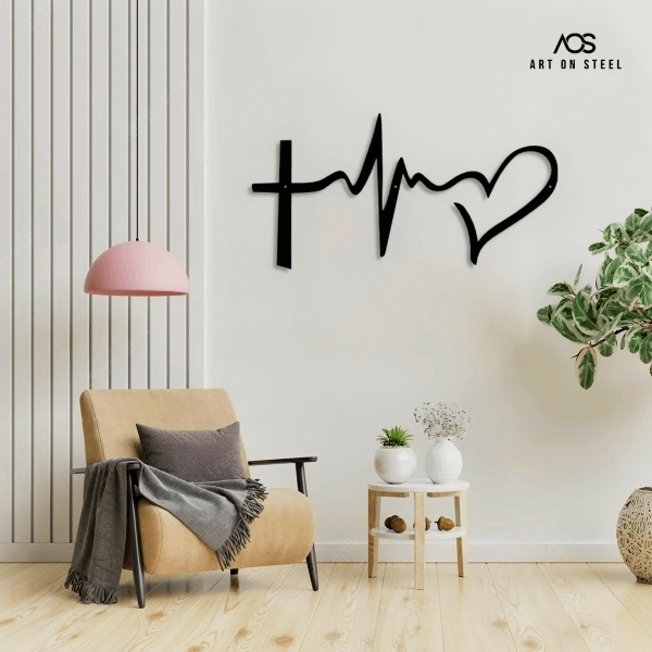 A large, black metal wall art piece depicting a cross, a heartbeat line, and a heart, symbolizing faith, hope, and love. It hangs above a modern beige armchair with a gray throw blanket and a small two-tier side table with decorative plants. A pink pendant light hangs in the left foreground. The "Art on Steel" logo is visible in the top right corner